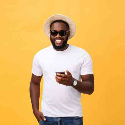 Cheerful African American man in white shirt using mobile phone application. happy dark skinned hipster guy read news from social network in mobile phone.